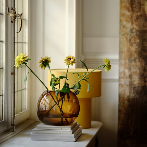Brun glasvas med gula blommor på bunten med böcker nära fönstret.