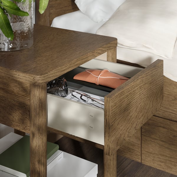 Sängbord i trä med öppen låda som avslöjar orange tyg, bredvid en säng med vita lakan.