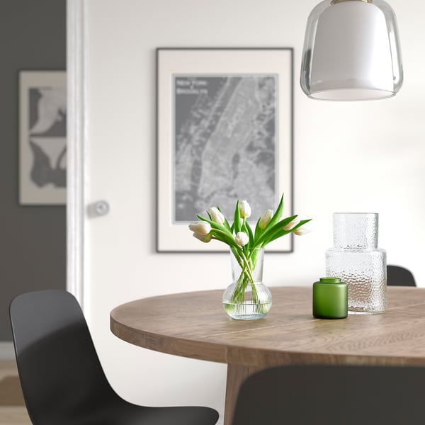 Ett matbord med en vas med vita tulpaner, ett grönt ljus och glasvaser. Konstverk och en grå taklampa hänger ovanför. Svarta stolar omger.