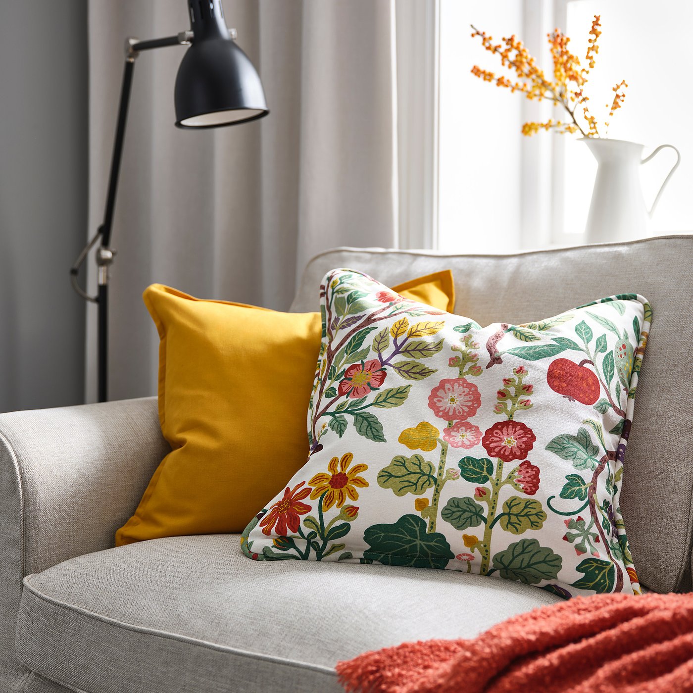 Mysig stol med blommig dyna VILDPERSILJA, större senapskudde, röd filt och orange blommor i en vit vas.
