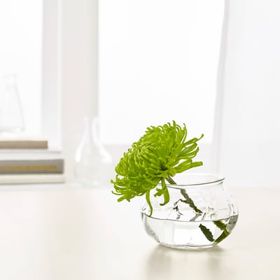 Den lilla glasvasen, VILJESTARK, rymmer en grön blomma. Elegant, rund design, sitter på det vita bordet.