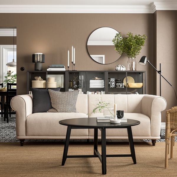 Ett mysigt vardagsrum med en VISKAFORS beige soffa, svart soffbord och prydnadsföremål på en mörk hylla mot en brun vägg.