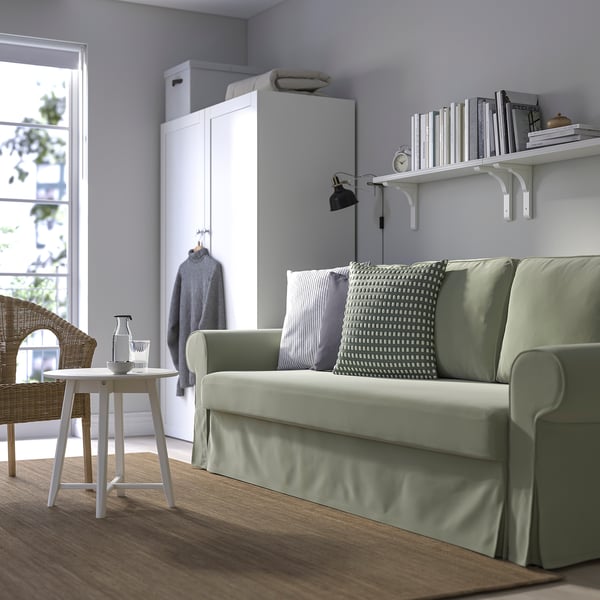 Grön VRETSTORP bäddsoffa i vardagsrummet, bredvid garderoben, under bokhyllan. Minimalistisk design med trägolv, vitt bord och stol.