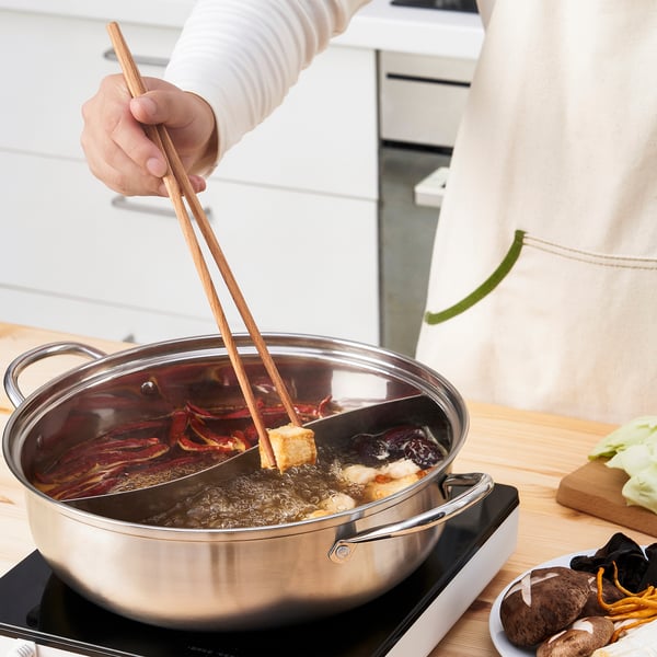 Person uses wooden chopsticks to cook tofu in a large pan with broth and vegetables on a stove.