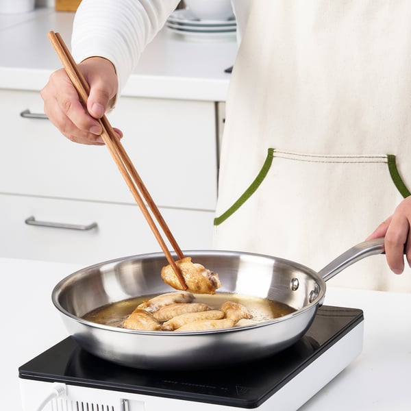 Person frying food with chopsticks in pan on modern stove, wearing long-sleeve shirt, in clean kitchen.