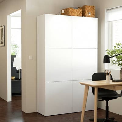 Large white BESTÅ cabinet stands tall near a window, taller than the desk beside it, with wicker baskets on top and sleek, minimalist design.