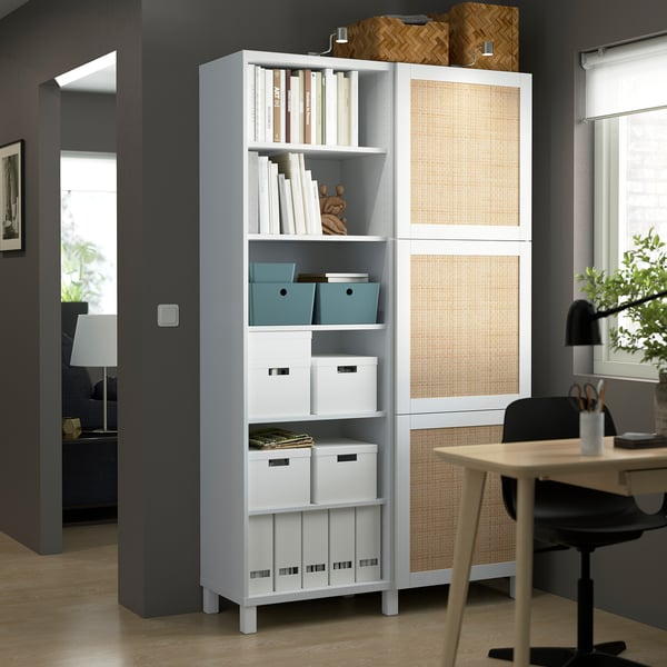 White BESTÅ storage unit with shelves, containing blue and white storage boxes and books. Its taller than the nearby desk and chair.