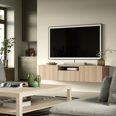 Modern living room with BESTÅ tv stand: light wood, minimalist design, large flat screen, plants, books, grey chair, natural light.