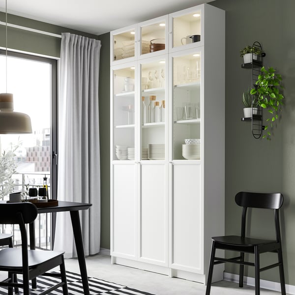 White BILLY bookcase with glass doors dishes and decor. Tall, narrow, fits neatly by window, surrounded by dark chairs.