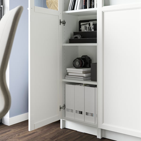 A white BILLY bookcase with glass panel; adjustable shelves hold books and a camera, while a cabinet below conceals clutter.