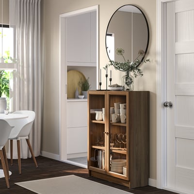 Modern living room with BILLY brown cabinet, glass doors, adjustable shelves, black mirror, and flower vase.
