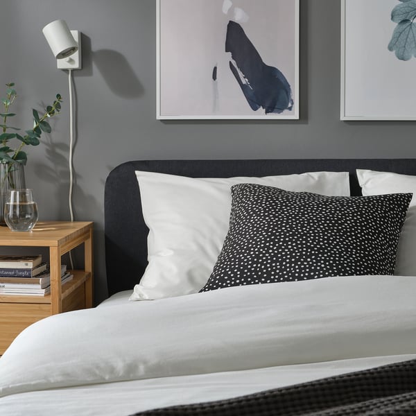 Modern bedroom with grey headboard, white pillows, dotted pattern cushion, wooden bedside table, books, plant vase, and wall art.