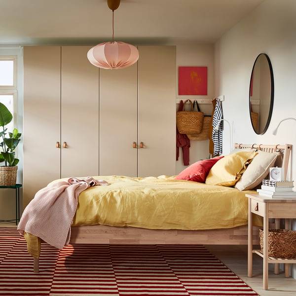 Cosy bedroom with beige walls, yellow bedding, red striped rug, hanging lamp, round mirror, plant, and wooden furniture.