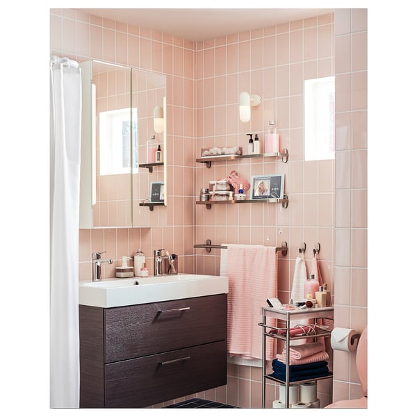 Modern bathroom with pink tiles, dark wood cabinet, white sink, wall shelves with bottles, towels, and decor, mirror with lights.