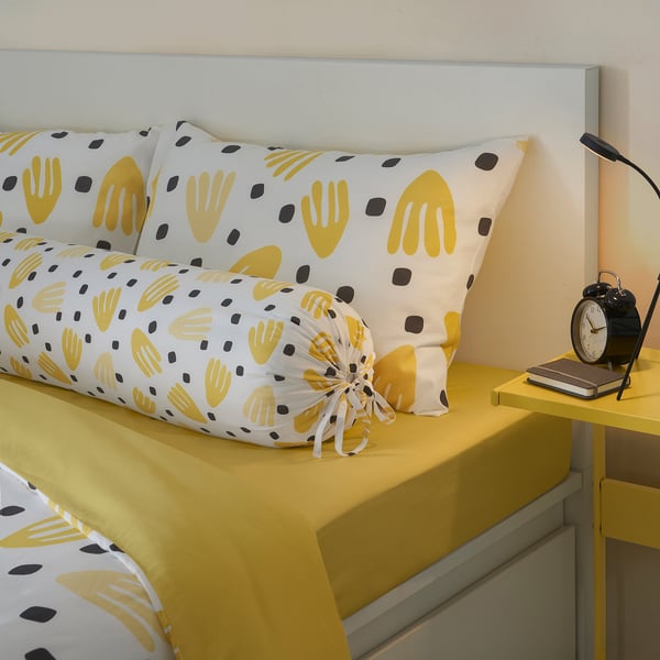 Bright yellow bedding with white hand designs. Sheets and pillows arranged neatly. Includes an alarm clock and book on a small bedside table.