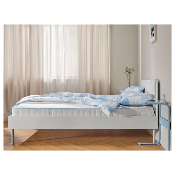 Modern bedroom with white and blue bedding on a firm mattress. Sheer curtains, wooden floor, side table with flowers.