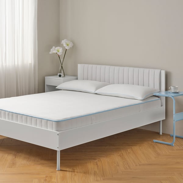 Modern minimalist bed setup. Large bed with headboard, wooden floor. Bedside table with flowers, blue chair with cup.