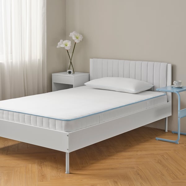 Minimalist bedroom with white BRUKSVARA mattress, bedside table, and blue chair. Firm, pocket spring design.