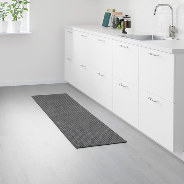 A small, rectangular kitchen mat named BRYNDUM in grey. It a non-slip backing and recycled polyester material, placed near white cabinets and a sink.