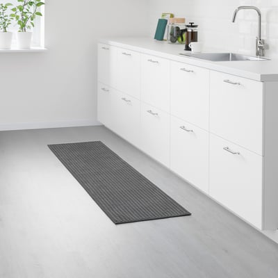 A small, rectangular kitchen mat named BRYNDUM in grey. It a non-slip backing and recycled polyester material, placed near white cabinets and a sink.