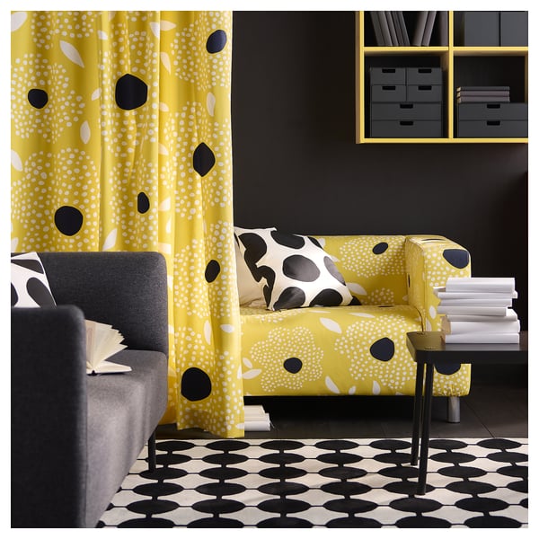 Bright living room with yellow curtain and matching sofa, black and white accessories, patterned rug, grey wall shelves, and open book on dark sofa.