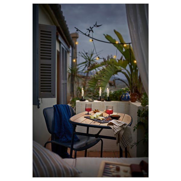 Small outdoor table, DUVSKÄR, wooden top, metal legs, foldable, set for two with wine glasses and food, surrounded by plants.