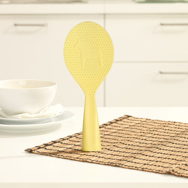 Yellow standing rice paddle on kitchen mat near plates and bowl.