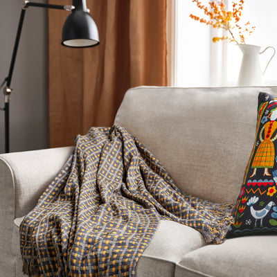 Cosy living room with grey couch, FLIKBRUNÖRT blanket, colourful pillow, orange flowers, black lamp.