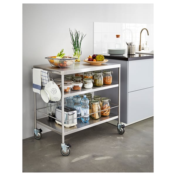 A silver FLYTTA kitchen cart with stainless steel shelves, lockable wheels, and various kitchen items including jars, vegetables, and kitchen tools.
