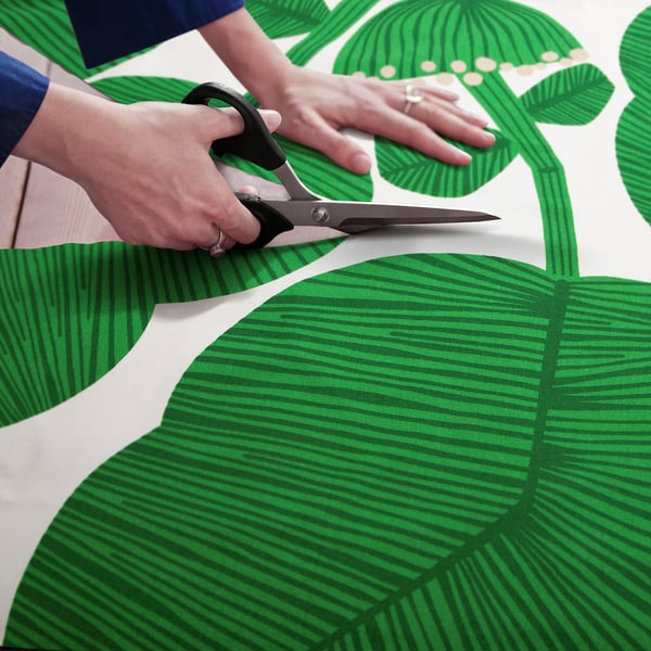 Person cuts green leaf-patterned fabric with scissors, preparing it for sewing projects like curtains or home decor.