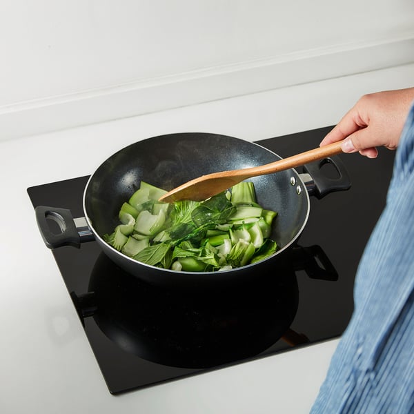 A person stir-fries vegetables in a two-handled non-stick kadai wok using a wooden spatula.