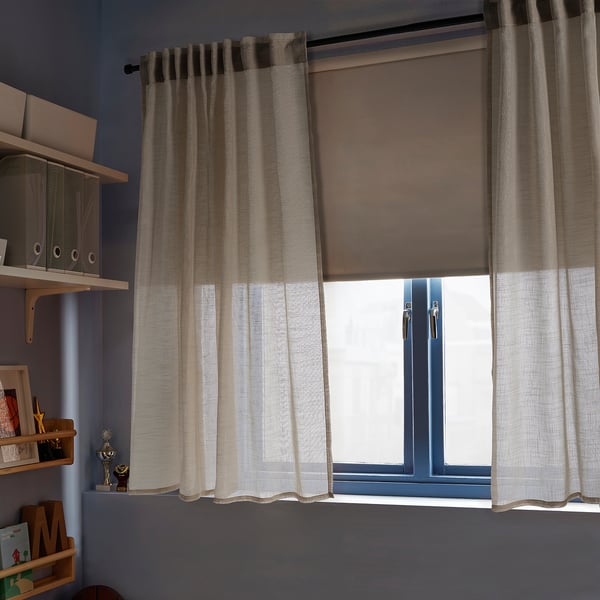 Beige sheer curtains with linen look hang from a wide window, covering a dark brown blind, flanked by wooden shelves.
