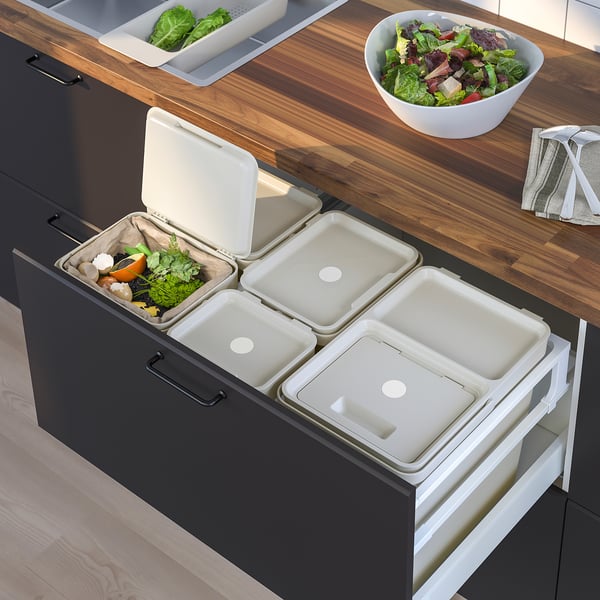 Kitchen setup showing HÅLLBAR waste sorting bins, dark grey, with lids open, containing miscellaneous waste. Wooden countertop with salad bowl.