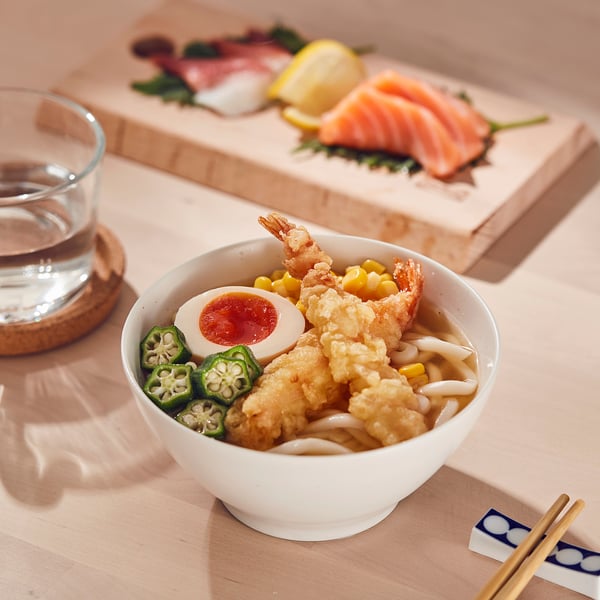 Bowl of udon noodles with tempura and vegetables. Chopsticks and sashimi platter nearby.