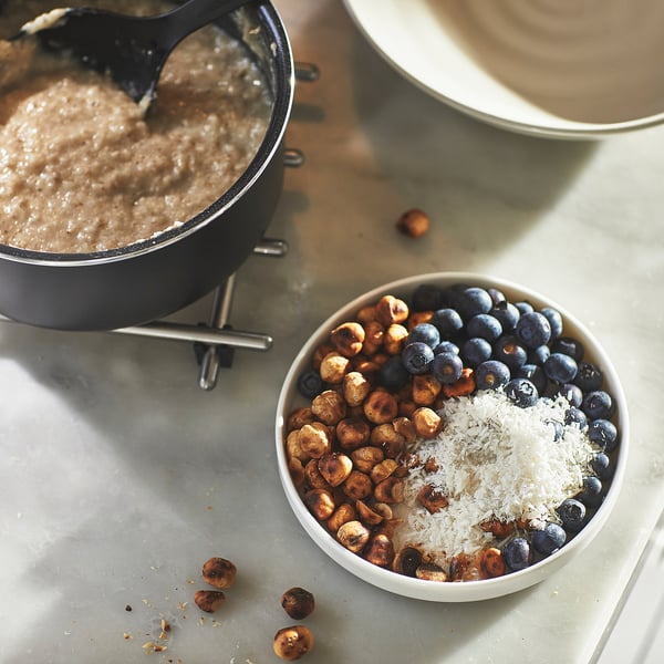 Small saucepan with non-stick coating, filled with a beige mixture, sits on a stand. Beside it, a bowl holds nuts, blueberries, and cheese.
