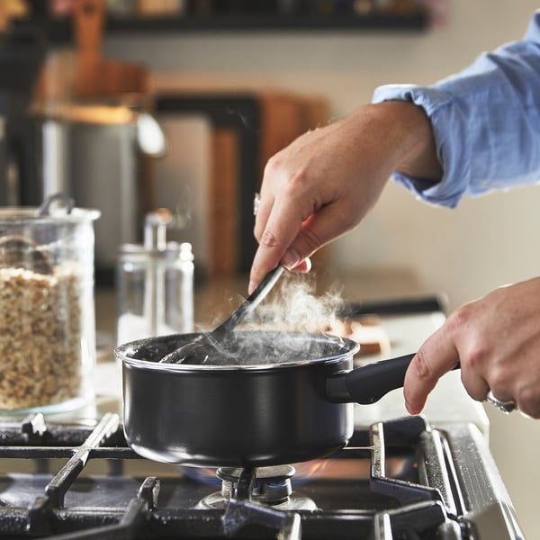 A person stirs food in a black non-stick saucepan on a gas stove with blue flames and rising steam.