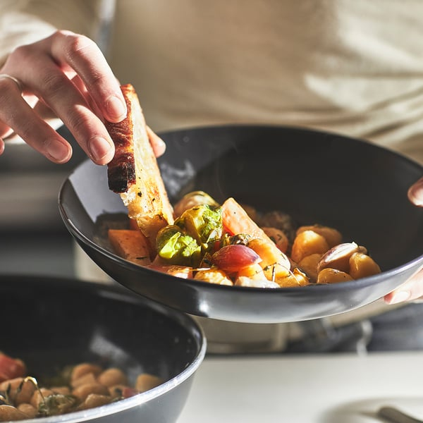 A hand holding bread dips into a bowl of stew, showcasing the HEMLAGAD pans even heat distribution and non-stick cooking surface.