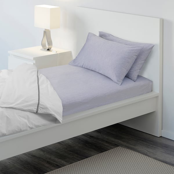 Bed with white headboard, light purple sheets, and a bedside table with a lamp.