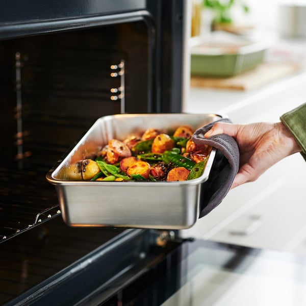 A hand holds a stainless-steel food container, filled with roasted vegetables, preparing to place it in an open oven.
