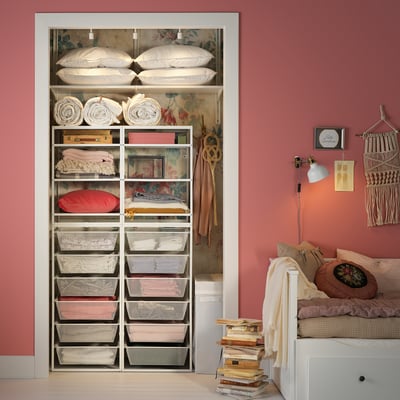 Organised closet with JONAXEL storage in pink room. Shelves hold folded linens, pillows, and mesh baskets. Coats hang on hooks. Bed and books nearby.