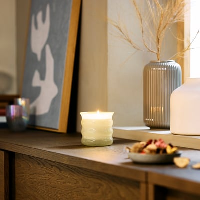 Cosy living room featuring a small, lit JUVELTRAST vanilla scented candle, wooden table with dried flowers, abstract art, and scattered fruits.