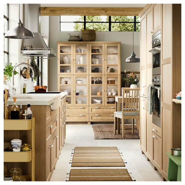 Modern kitchen with light wood cabinets, island, and glass-door display unit. Stainless steel hood, pendant lights, and a dining table with chairs.