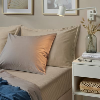 Cosy bedroom with beige LÄKEVÄNDEROT sheet, peach pillow, bedside table with book and flowers, wall art, and reading light.