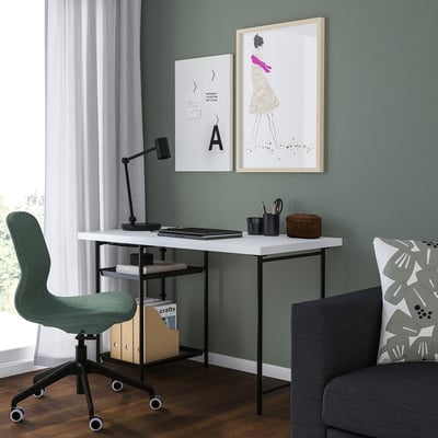 Minimalist home office with white desk, green chair, grey armchair, two artworks, black lamp, wooden floor, natural light.
