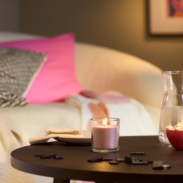 A cosy bedroom with a lit pink LOTSFÅGEL scented candle on a dark wooden table, near remote controls and popcorn. The candle is small.