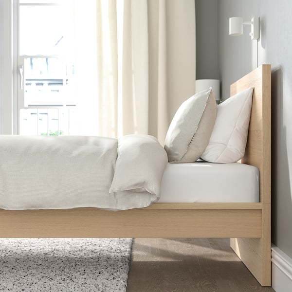 Minimalist bed with light wood headboard, white bedding, and striped pillows. Bright, airy room with natural light and city view.