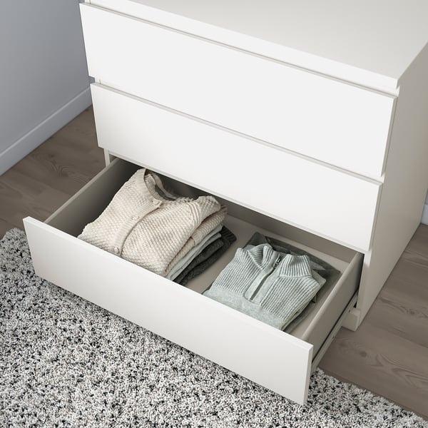 A white MALM chest of drawers with a partially open drawer holding neatly folded clothes, illustrating its storage function.