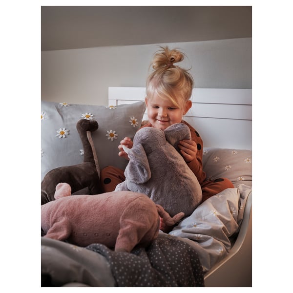 A young child sits on a bed holding a grey stuffed elephant cuddly toy. The bed has a beige comforter with white daisy designs.