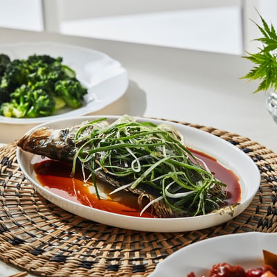 Platter holds cooked fish with green onions atop sauce, flanked by veggie side dishes, reflecting OFTASTs versatile modern serving style.