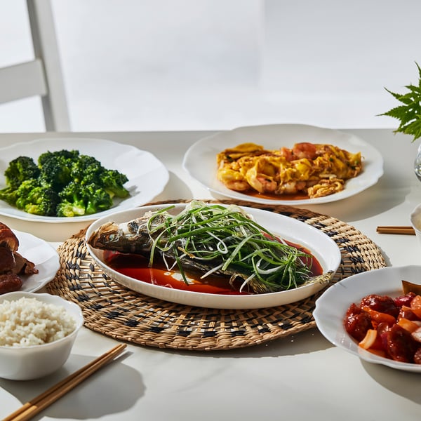 Five plates with broccoli, noodles, stew, fish, and rice. Chopsticks on a woven mat.